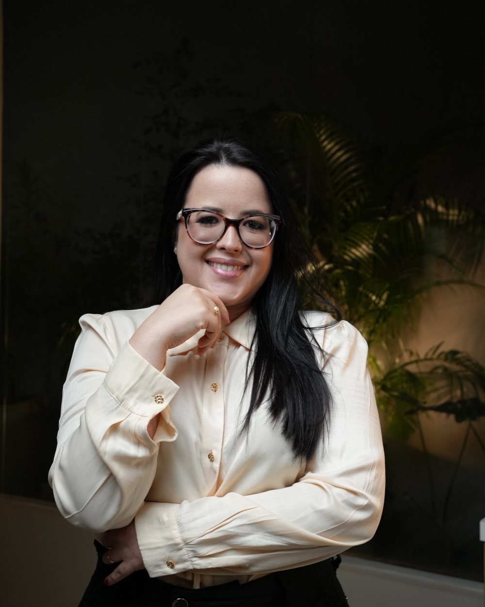 Mônica Mariane, psicóloga e neuropsicóloga, sorrindo com blusa clara em ambiente com plantas